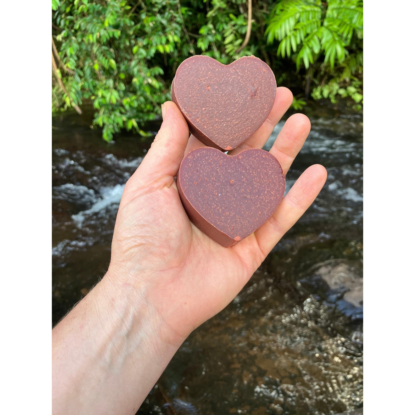 Hand holding two deep red heart soap against a natural creek water background, at a distance.