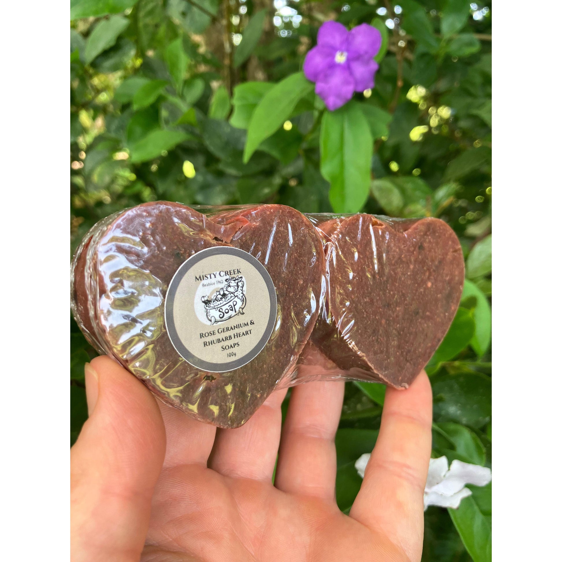Hand holding two clear packaged heart soaps with a green foliage natural background.