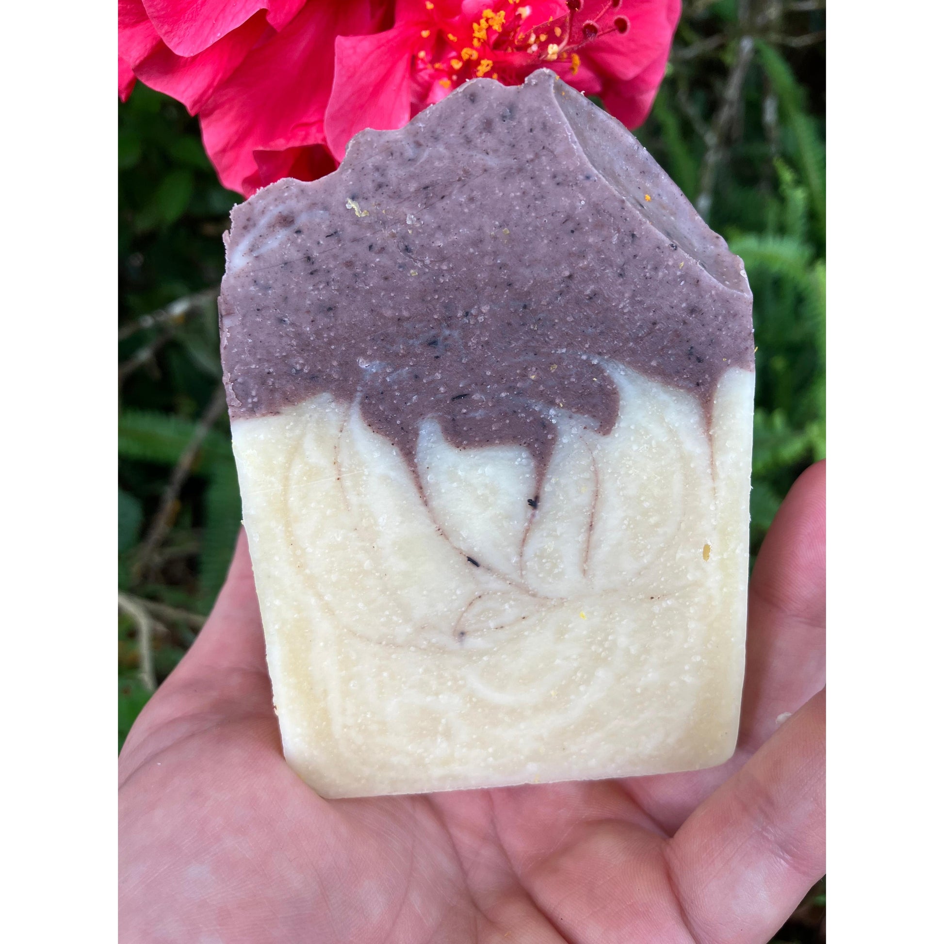 Hand holding a two-tone deep purple l organic lavender soap bar with pink flower in the background