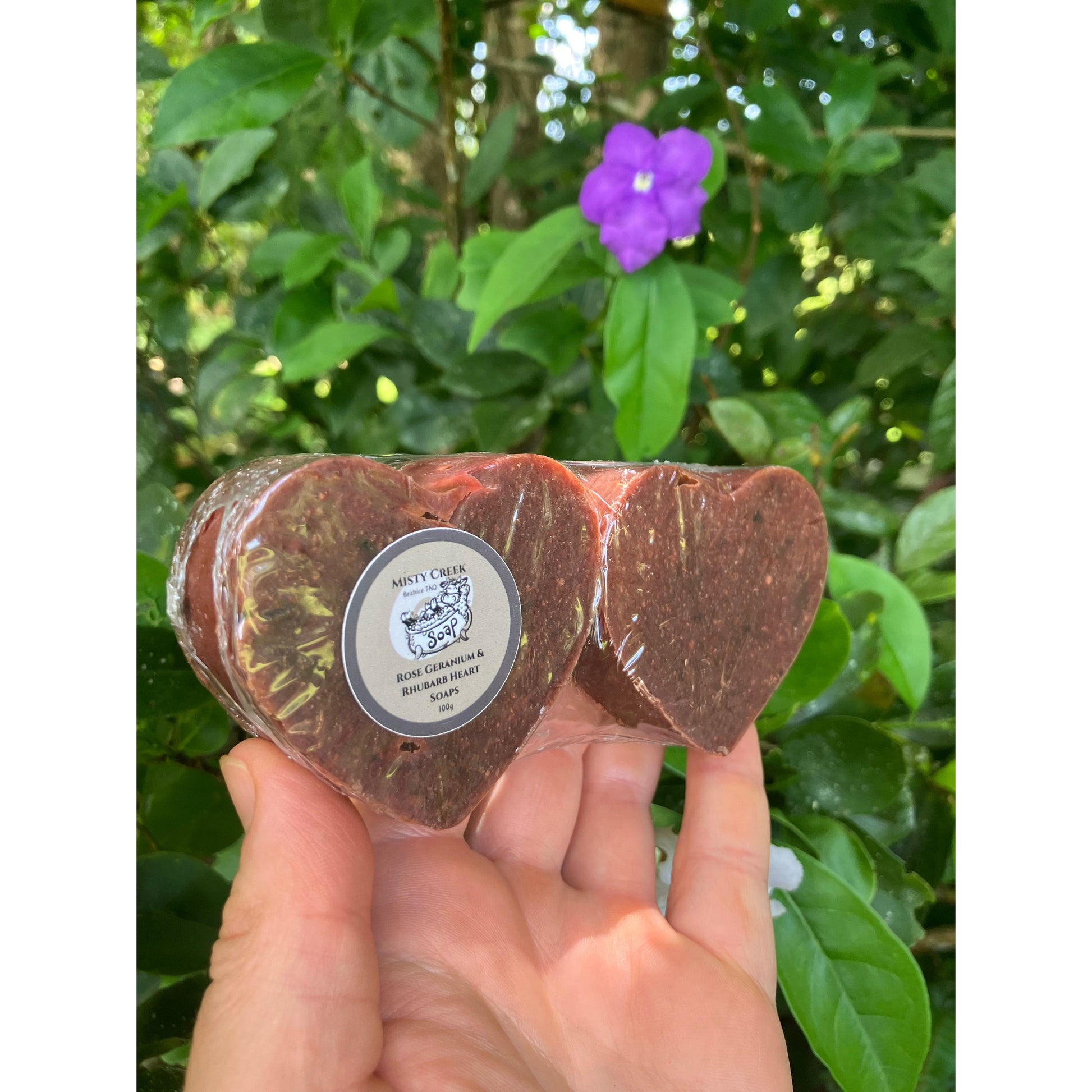 Hand holding two deep red heart soaps side by side in clear packaging with a green foliage background with a purple flower.