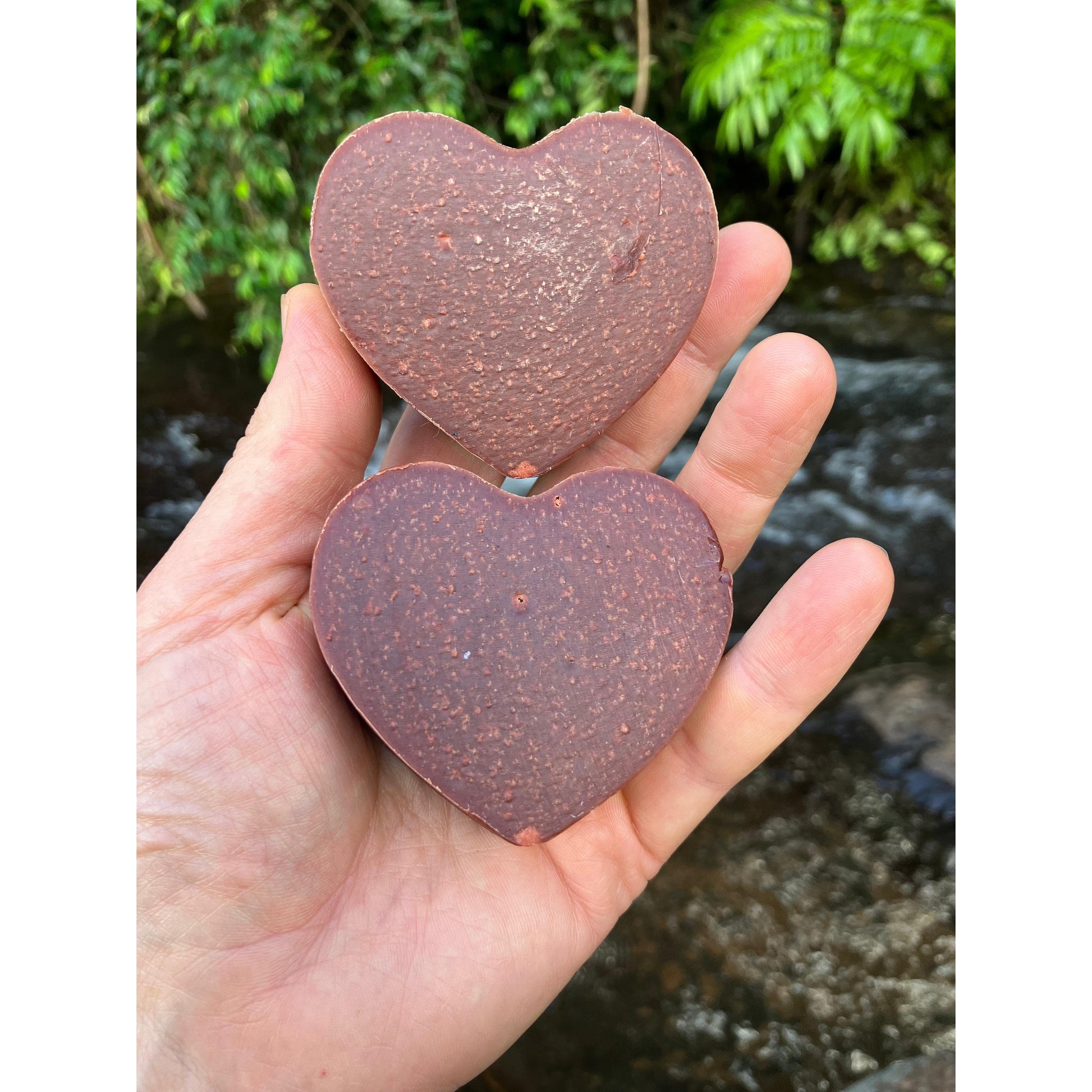 Hand holding two deep red heart soap against a natural creek water background.