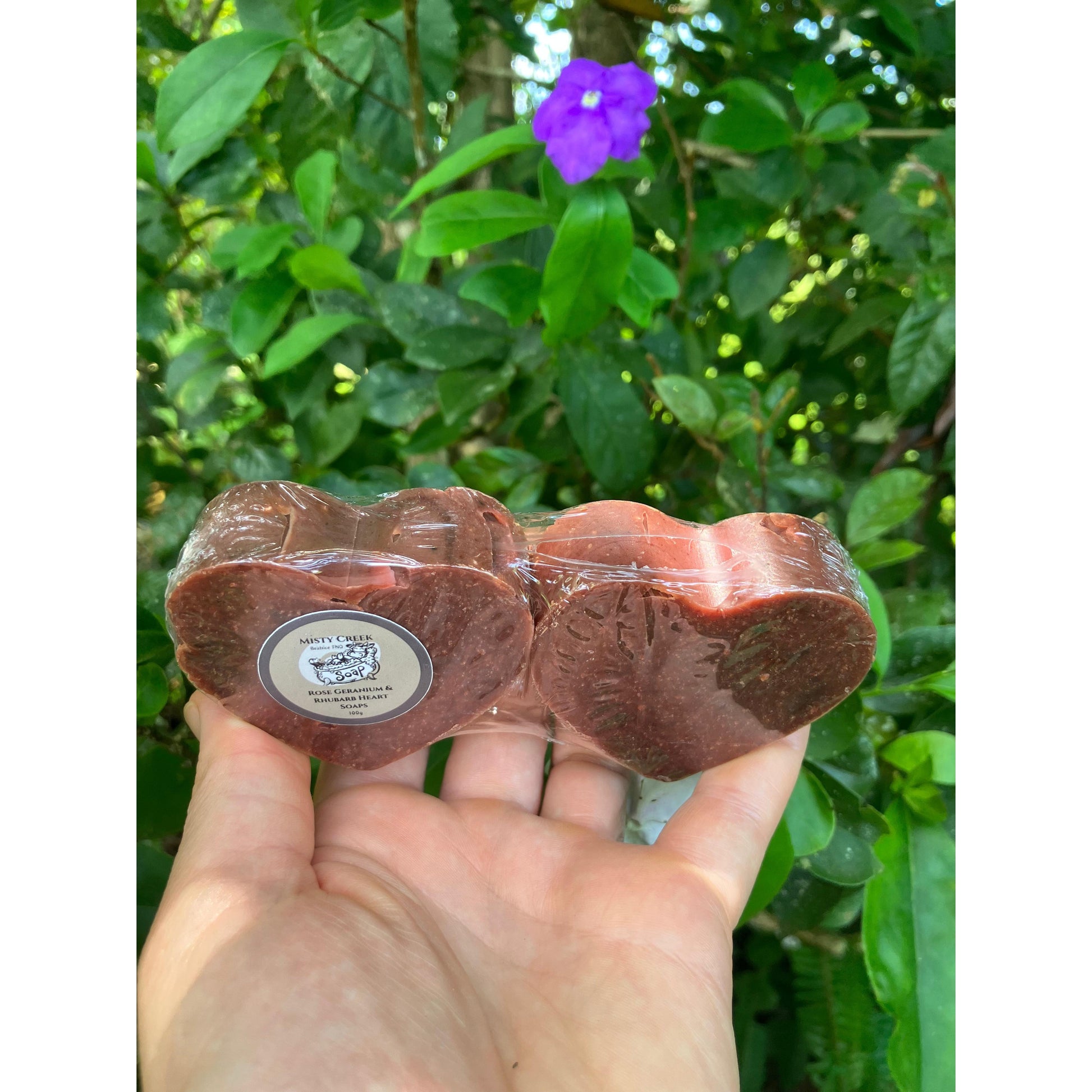 Hand holding two twin heart soaps in clear packaging by Misty Creek Soap against a green natural foliage background.